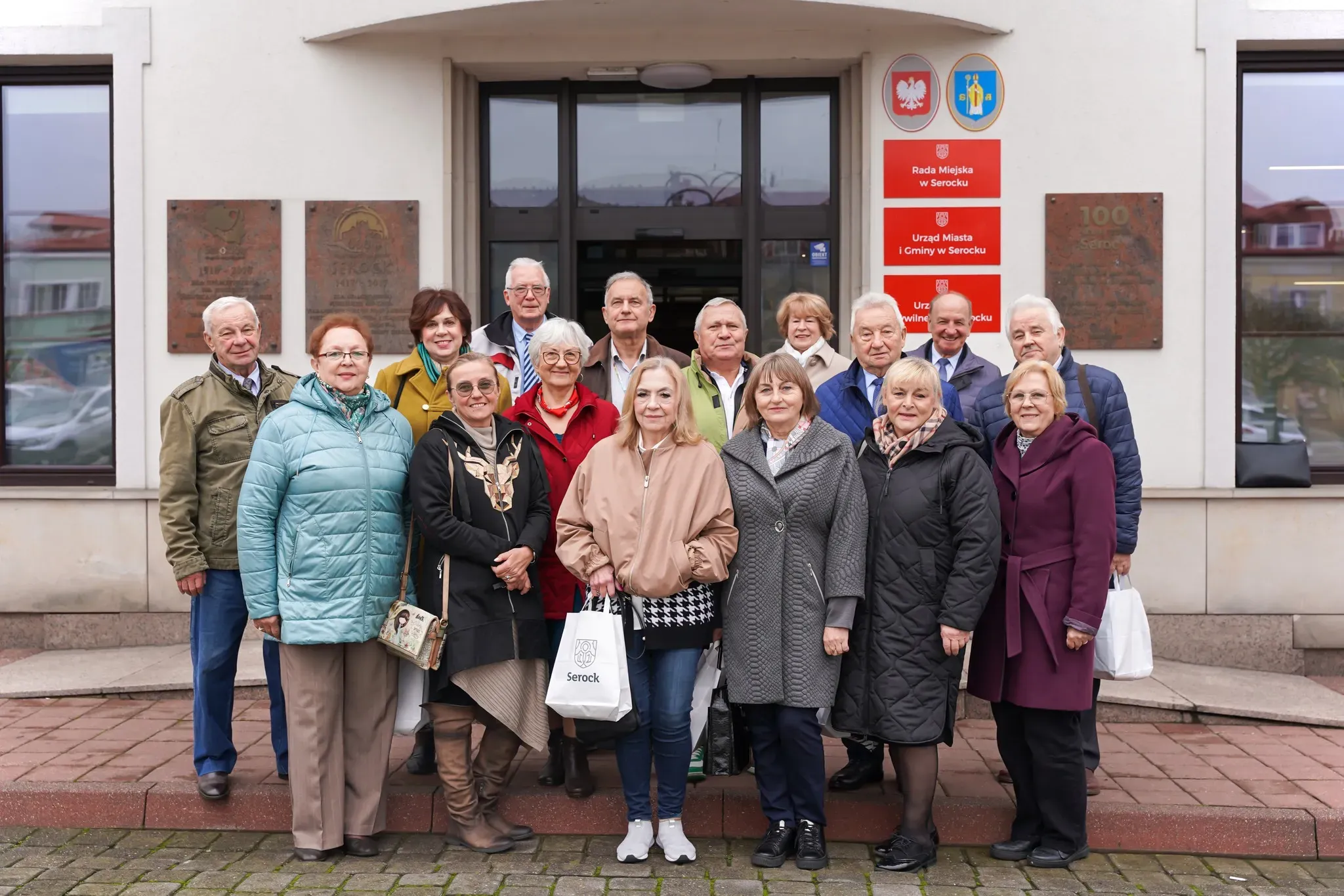 Na zewnątrz, w dzień grupa szesnastu osób stoi przed wejściem do budynku urzędu miasta. Większość osób ubrana w kurtki i płaszcze. W tle widoczne tablice z nazwami urzędu i herbem miasta.